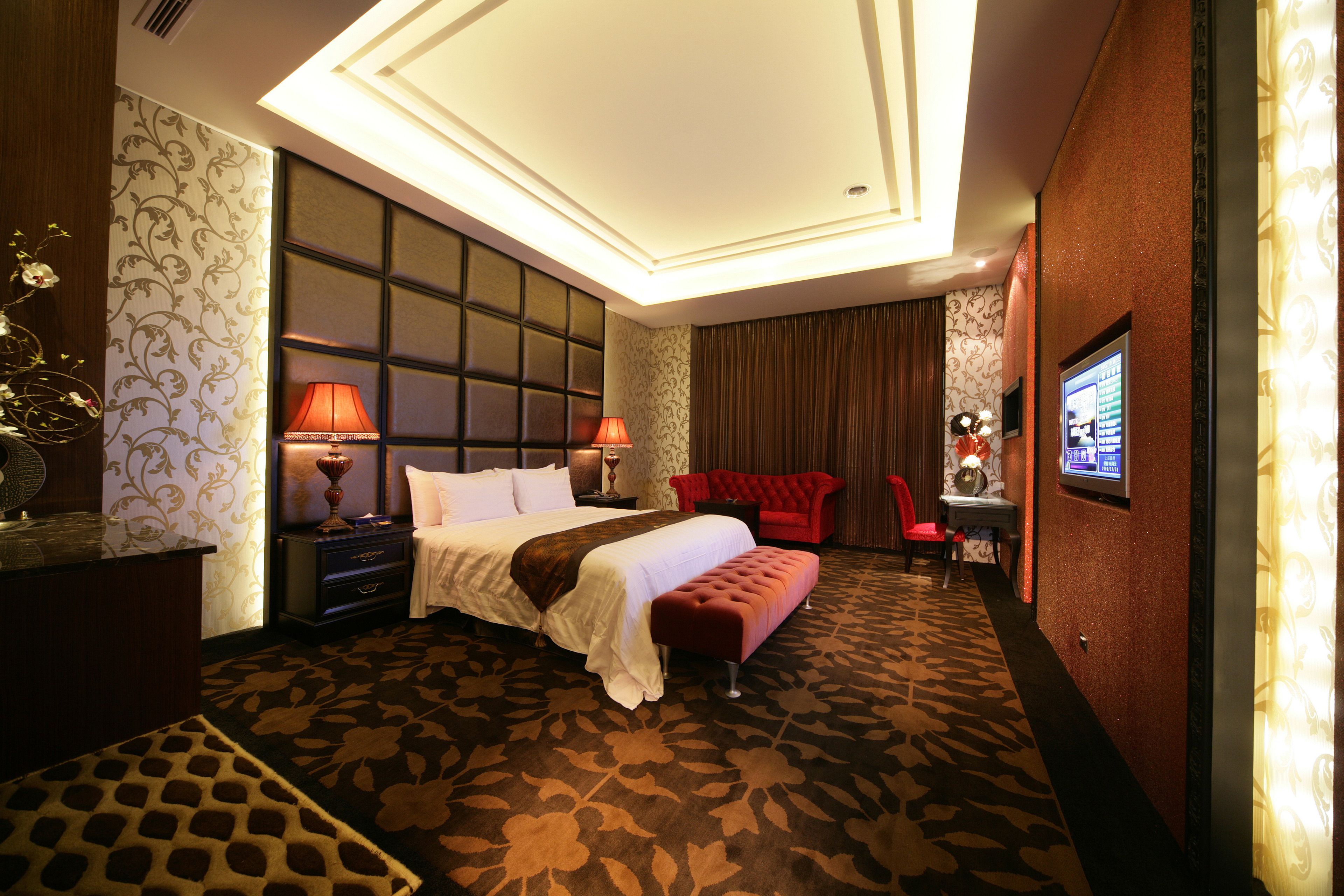 A hotel room with a large bed, two bedside lamps, a red armchair, a small table with a lamp, and a television on the wall.