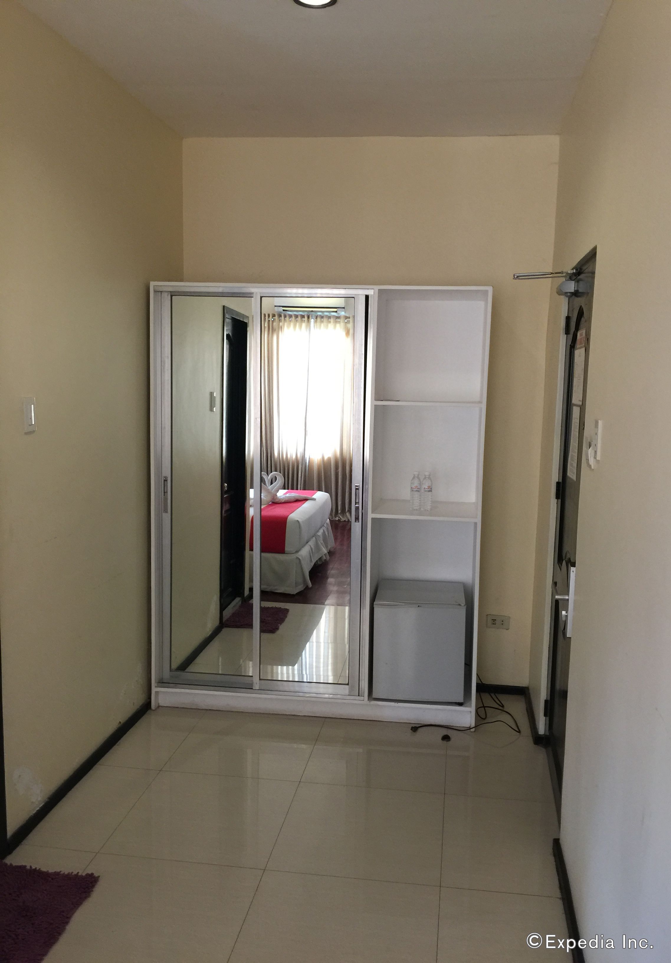 A hotel room with a sliding door closet, a bed with a red bedspread, and a small white cabinet.