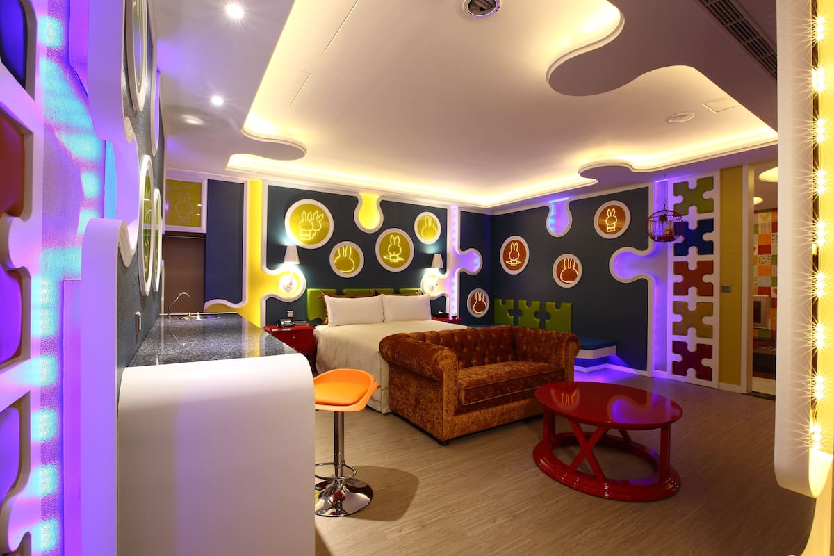 Une chambre d’hôtel moderne avec un grand lit, un canapé, un fauteuil rouge et une salle de bain équipée d’un lavabo et d’un miroir.