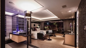 Una habitación de hotel moderna con una cama grande, una zona de estar con sofá y mesa de centro, una pequeña cocina y un baño con bañera y ducha.