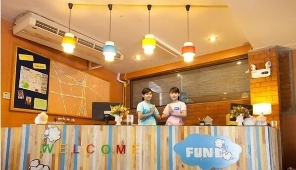 A reception desk with a 'WELCOME' sign and a 'FUND' logo, two staff members behind it, and a bulletin board with papers. - FUN-D Hostel Chiang rai (Chiang Rai)