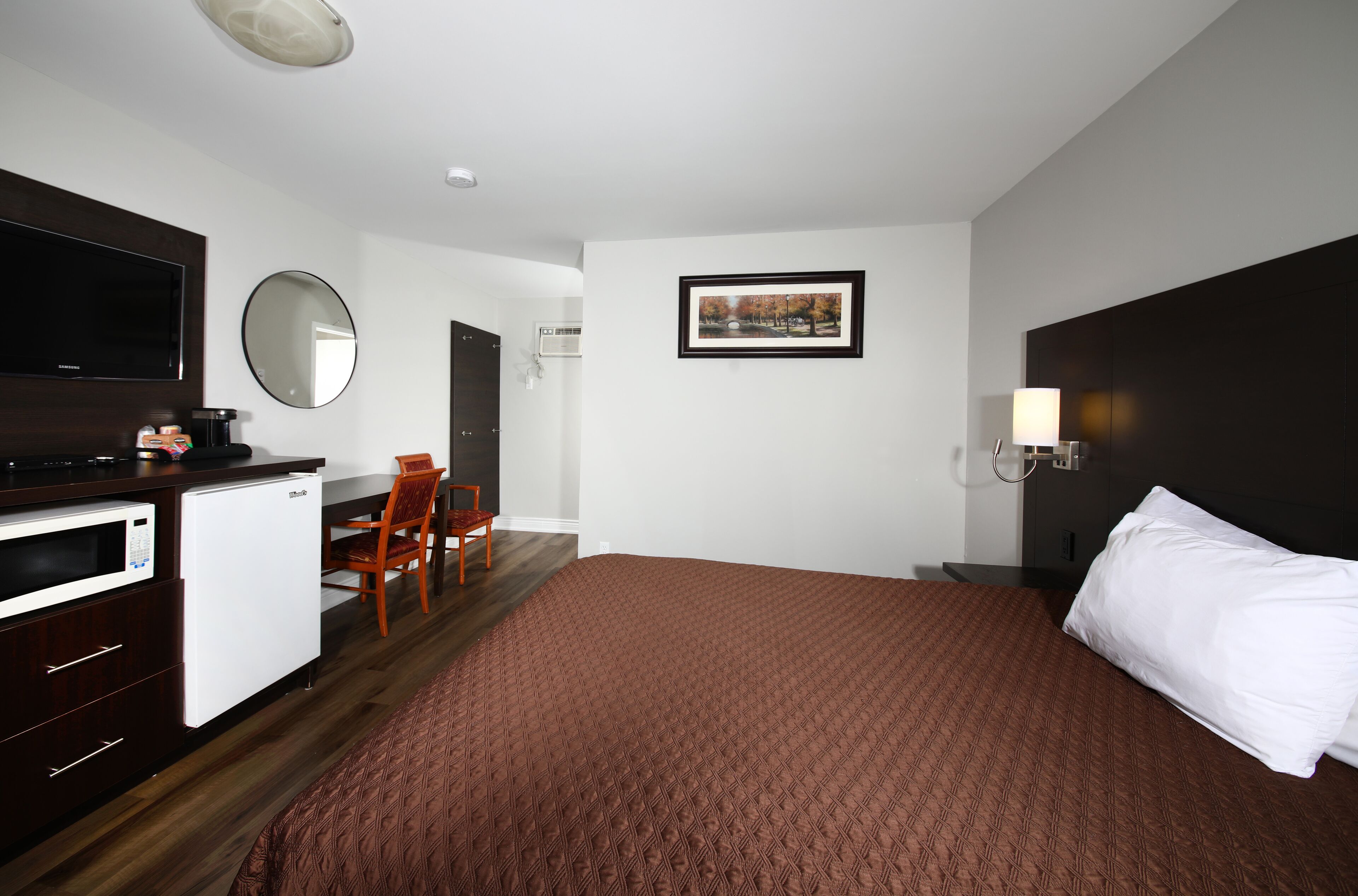 A hotel room with a bed, a desk with a microwave, a chair, a TV, and a framed picture on the wall.