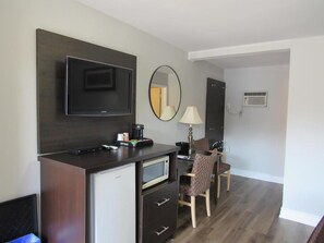 A compact hotel room with a kitchenette, a flat-screen TV, a mirror, a desk with a chair, and a small refrigerator.