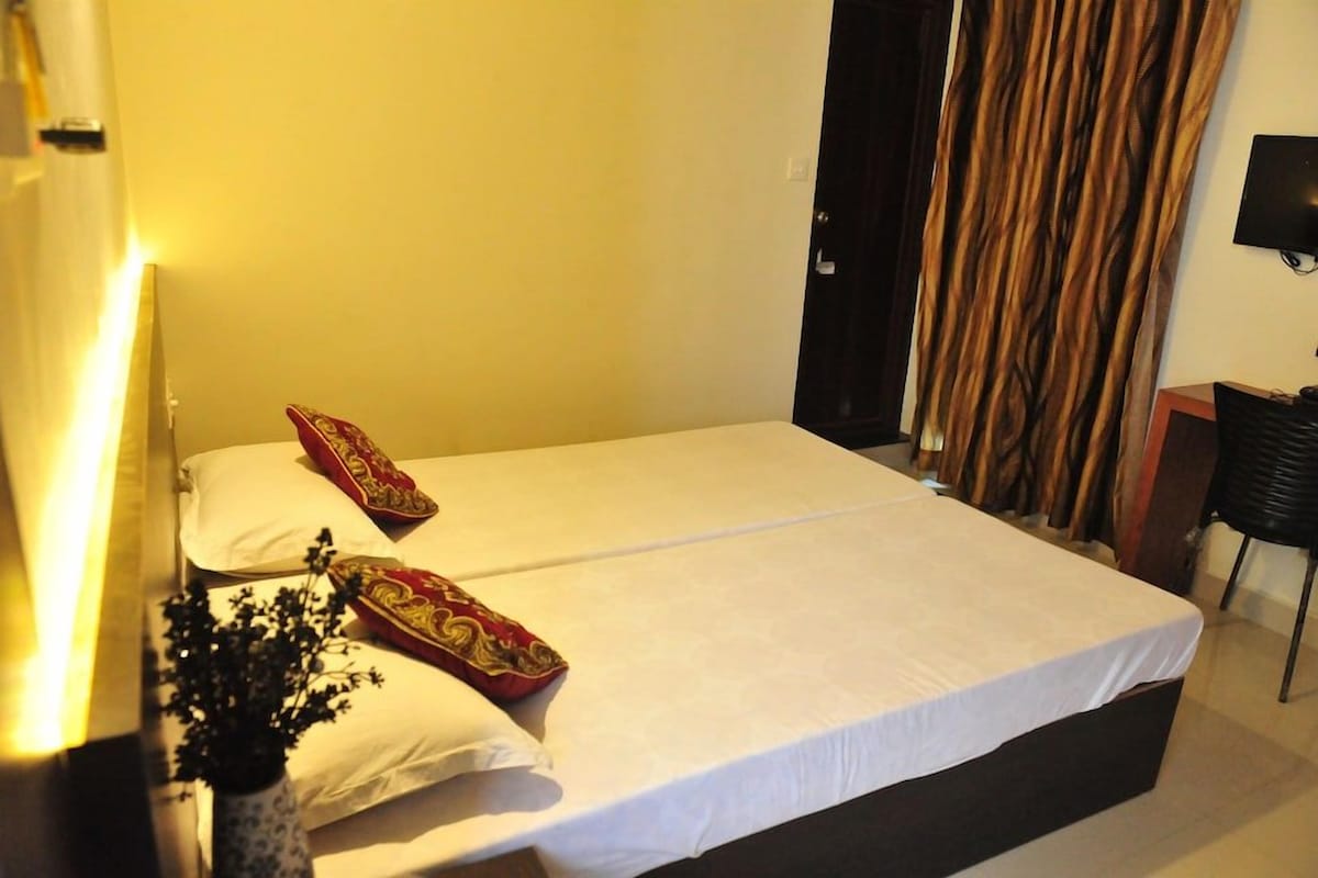 Un lit simple avec une literie blanche et deux coussins décoratifs, une table de chevet avec une lampe et un téléviseur fixé au mur.