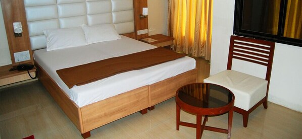 A hotel room with a large bed, a wooden nightstand, a bedside table, a chair with a cushion, and a small round table. - Hotel Aishwarya Residency (Navi Mumbai)
