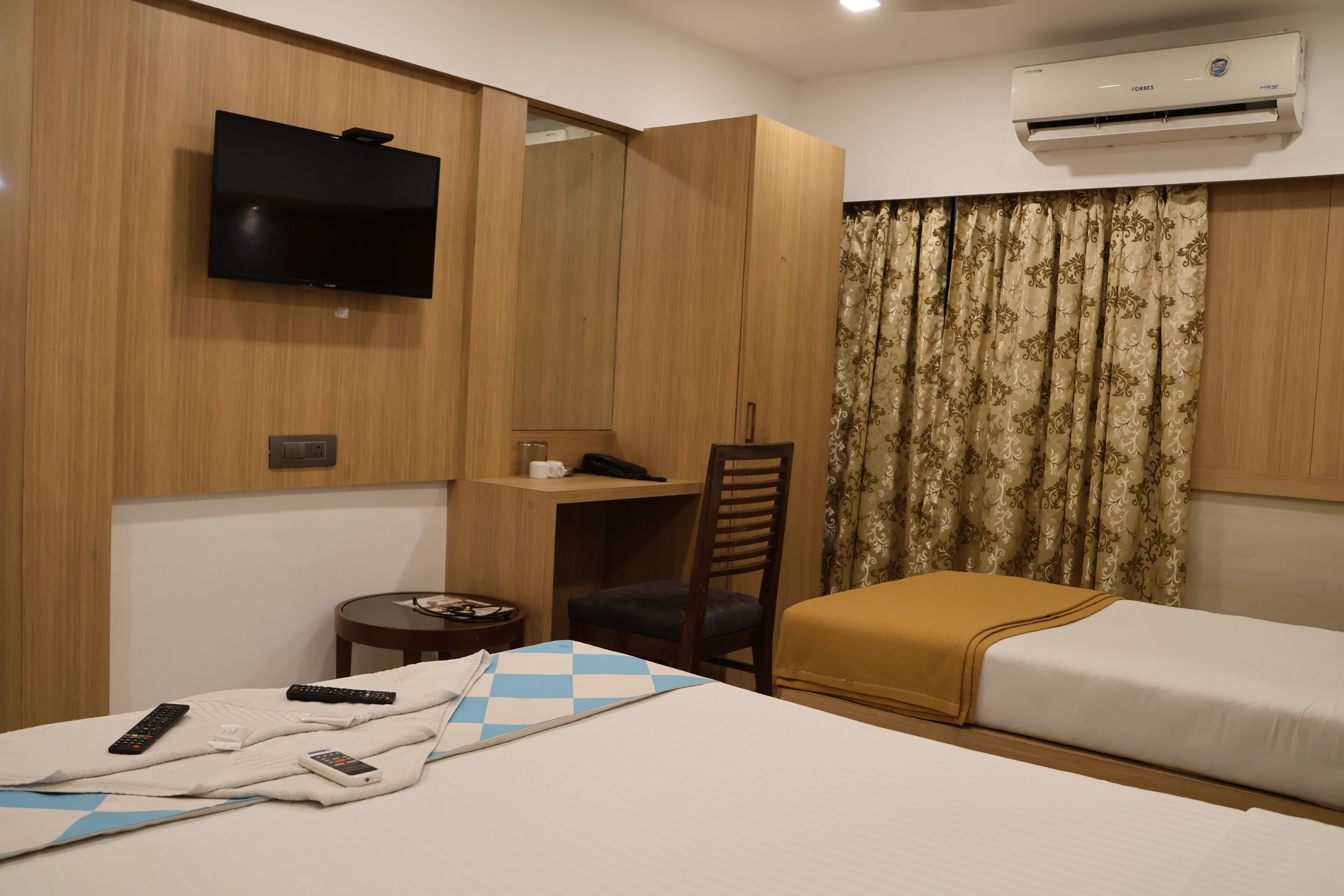 A hotel room with a bed, a desk, a chair, a TV, and an air conditioning unit.