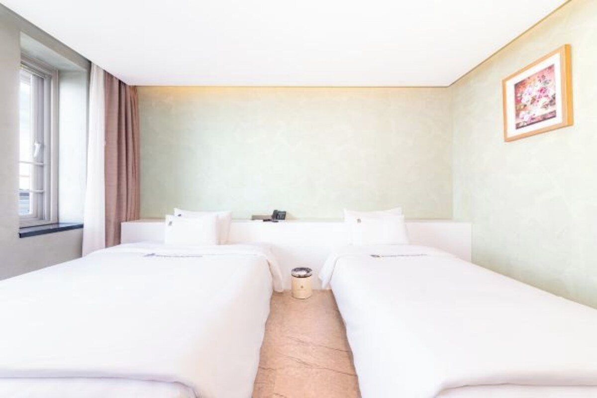 A hotel room with two single beds, a small bedside table, a nightstand, a window with curtains, and a framed picture on the wall.