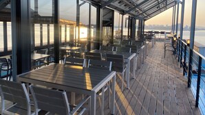 A covered outdoor dining area with tables and chairs, large windows, and a view of the water. - Benikea The Bliss Hotel (Incheon)