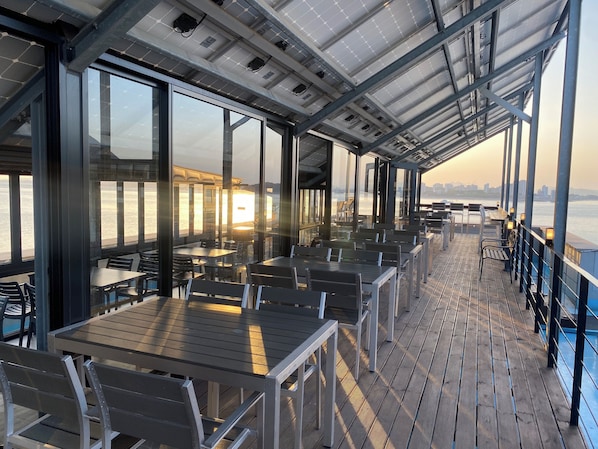 A covered outdoor dining area with tables and chairs, large windows, and a view of the water. - Benikea The Bliss Hotel (Incheon)