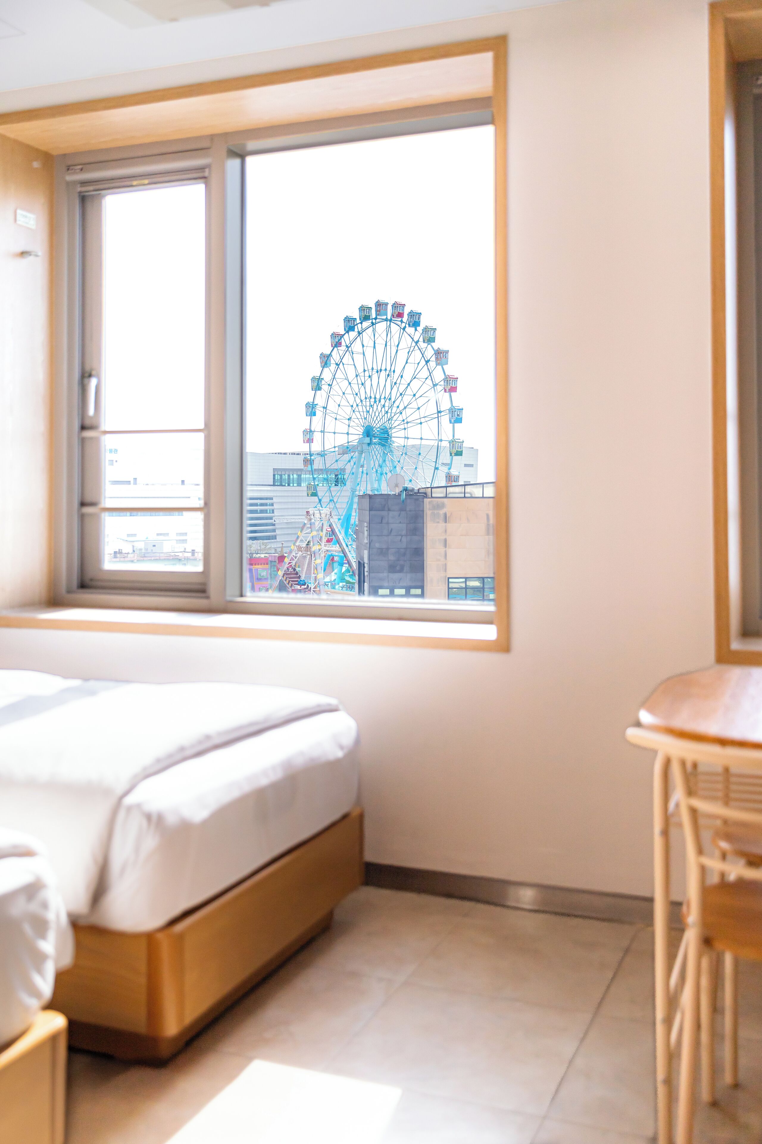 A hotel room with a bed, a wooden table, and a large window offering a view of a Ferris wheel.