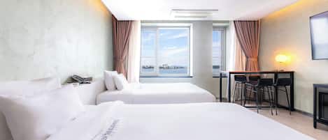 A hotel room with two beds, a desk with chairs, a large window with curtains, and a wall-mounted lamp.