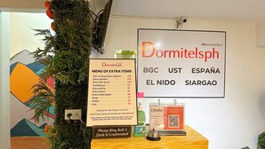 Un mostrador de recepción de hotel con un cartel que detalla artículos adicionales y sus precios, un timbre y un código QR para el registro en línea.