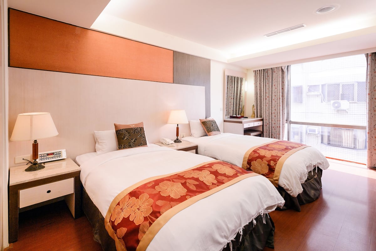 A hotel room with two beds, a nightstand with a lamp, a telephone, and a window with curtains.