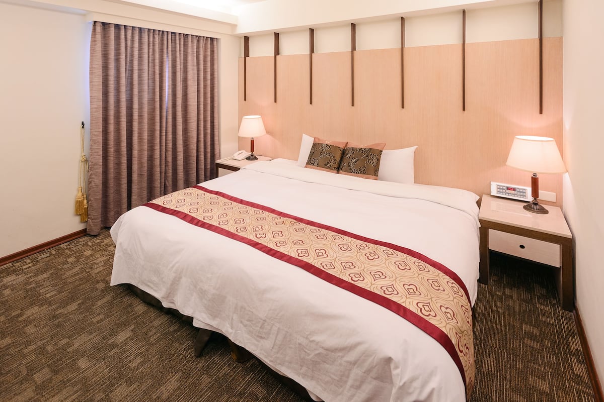 A hotel room with a large bed, two bedside tables, a headboard, and a curtain.