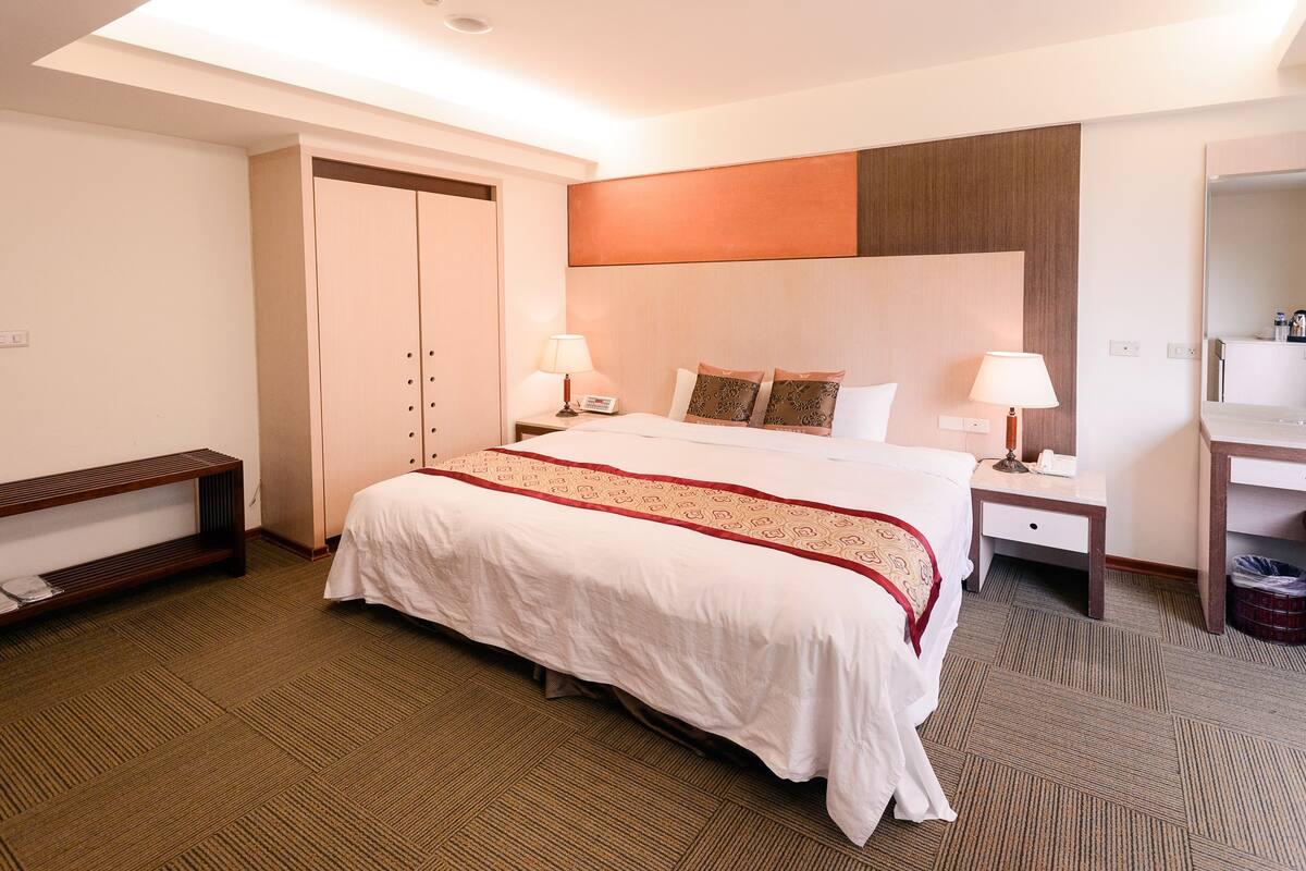 A hotel room with a large bed, two bedside tables with lamps, a desk, and a wardrobe.