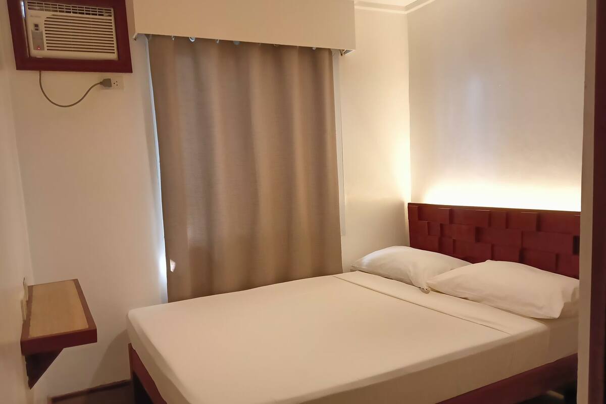 Een hotelkamer met een bed, nachtkastje, een wandairconditioner en een raam met gordijnen.
