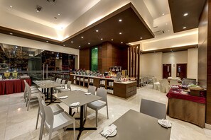 A modern dining area with multiple tables and chairs, a buffet setup, and a large window with a view of trees.