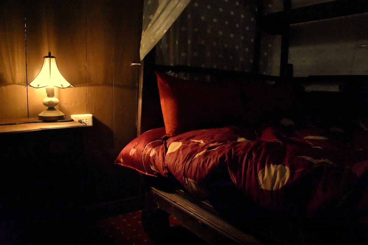 A dimly lit room with a wooden headboard, a single red pillow, and a table lamp on a wooden nightstand.