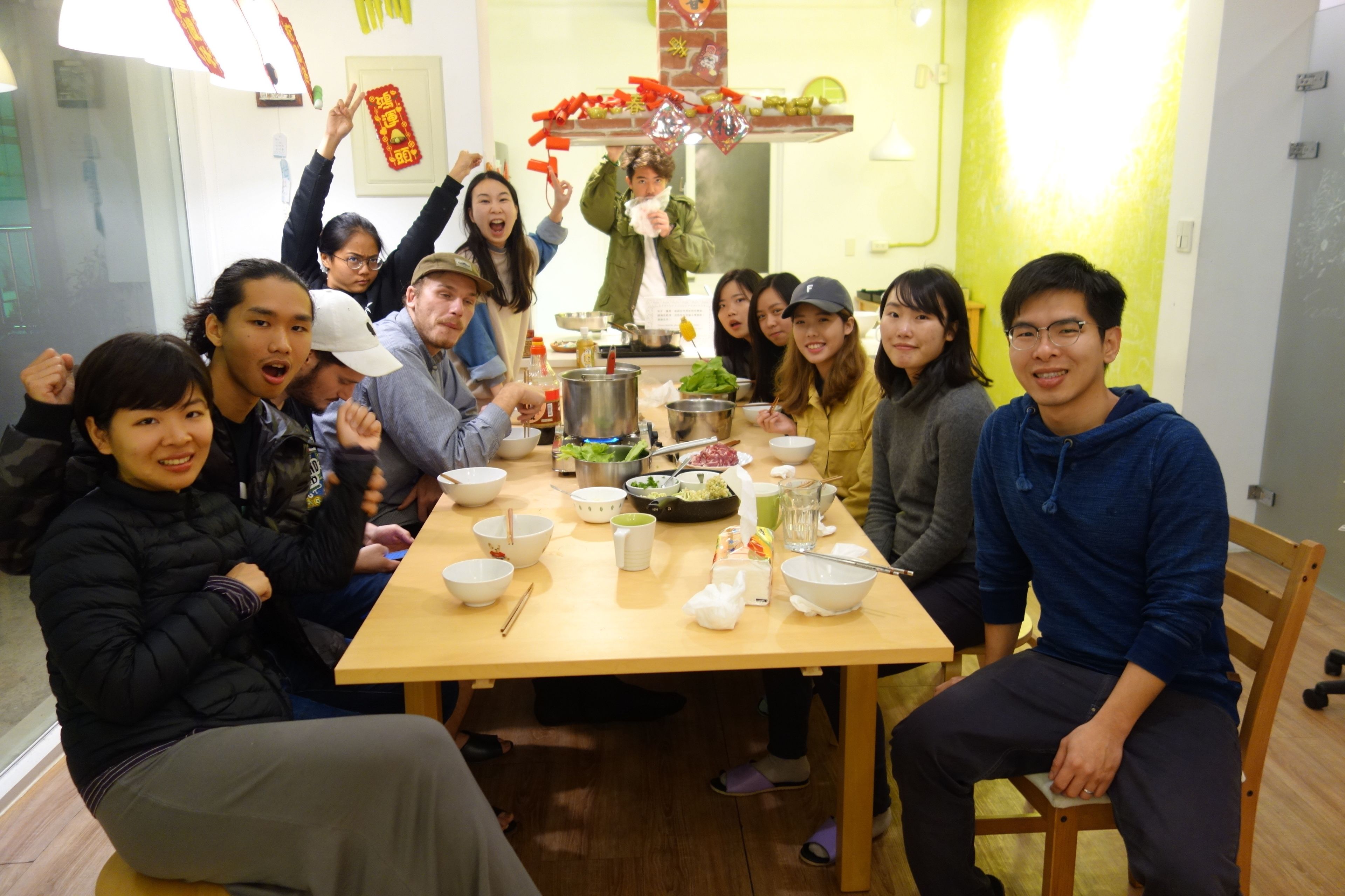 A group of people sitting around a table with various dishes and utensils, in a room with a wooden floor and a green wall.