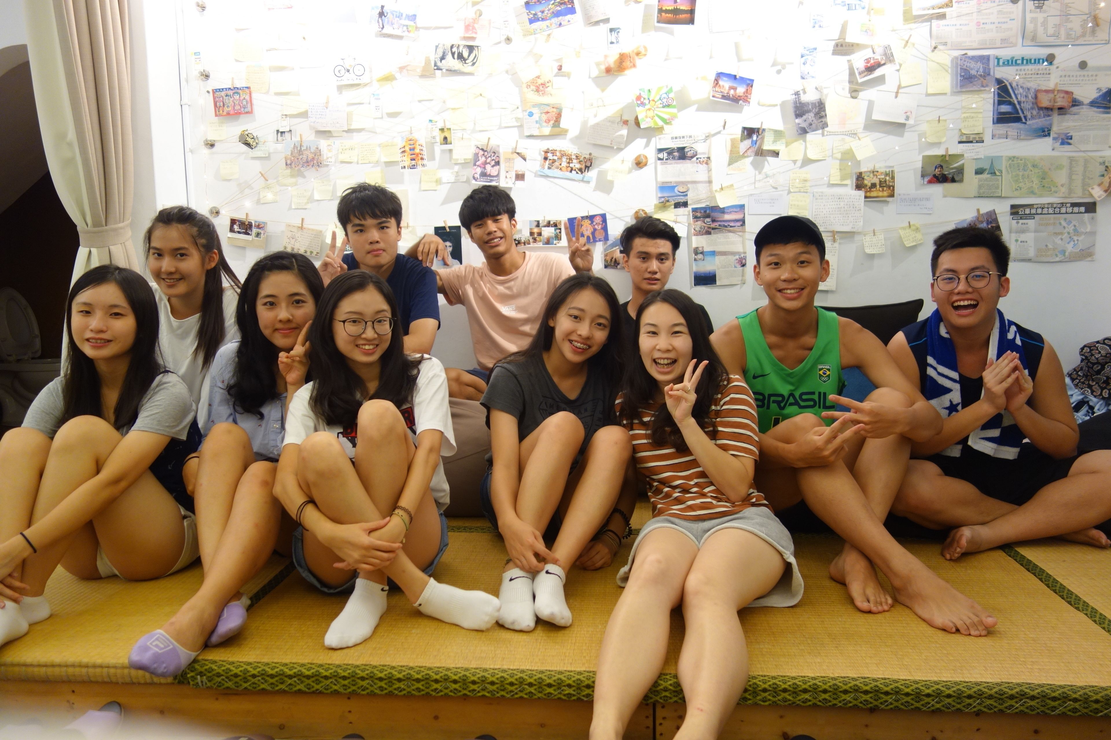 A group of people sitting on a tatami mat in a room with a wall covered in various papers and photos.