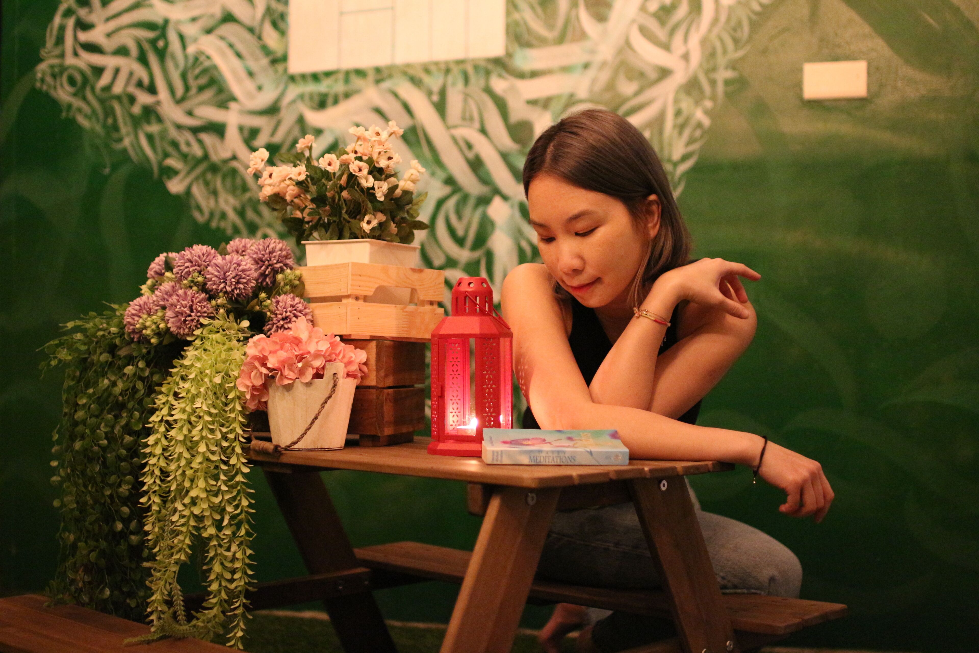 A person sitting at a wooden table with a red lantern, a book, and various flowers.