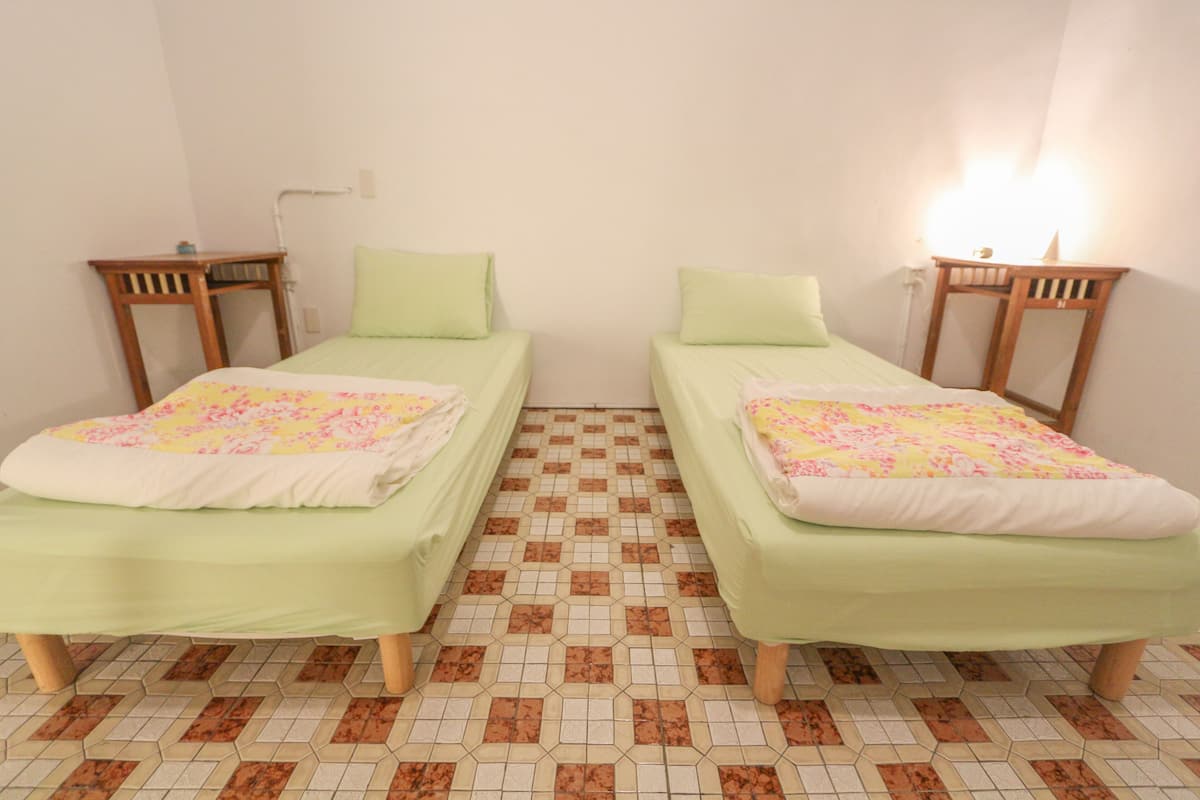 Two single beds with green bedding and floral patterns, a tiled floor with a geometric pattern, and bedside tables with lamps.