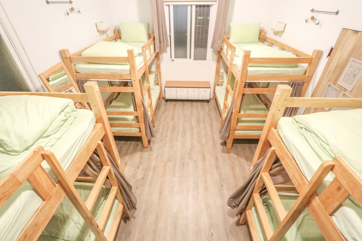 A dormitory room with bunk beds arranged in a U-shape, a window with curtains, and a small bench in the center.