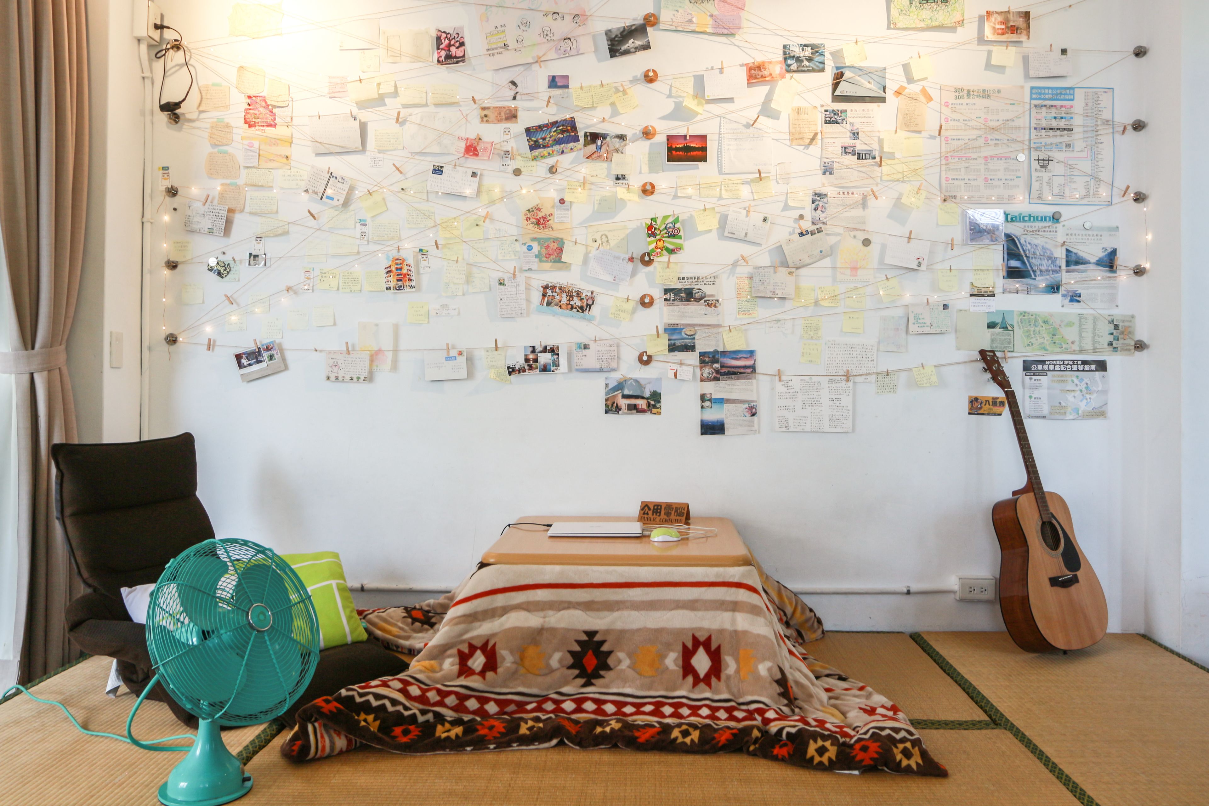 A room with a bed, a fan, a guitar, and a wall covered with notes and photos.
