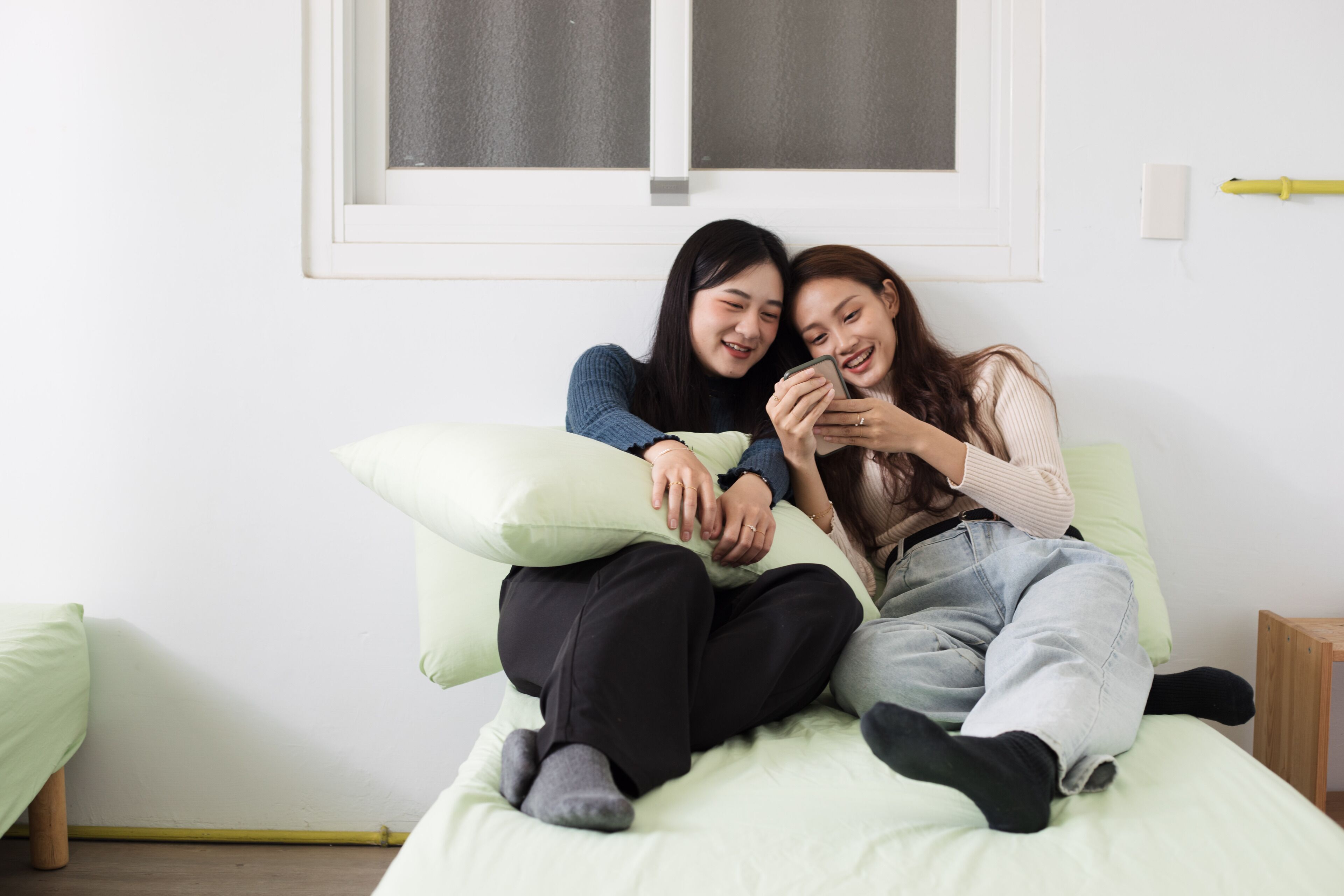 Twee personen zitten op een bed met kussens, in een kamer met een raam en een klein houten krukje.