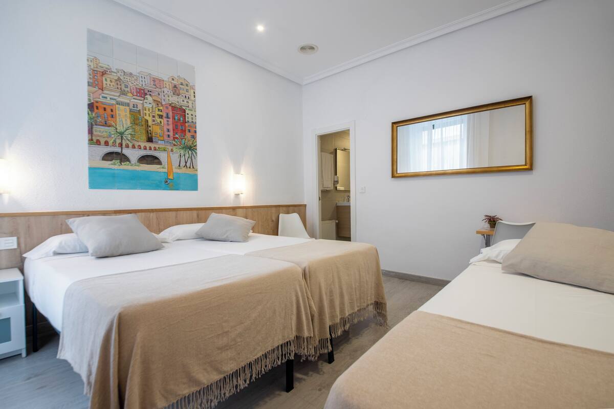 A hotel room with two beds, a large painting of a coastal town, a mirror, and a nightstand.