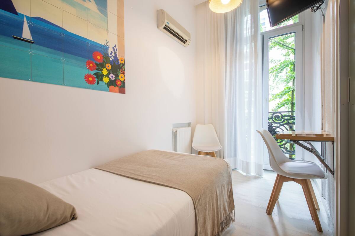 A hotel room with a bed, a desk with a chair, a TV, an air conditioner, and a window with a view of greenery.