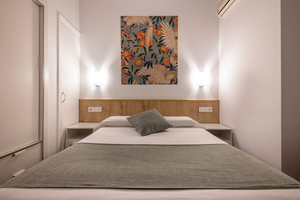 A hotel room with a bed, a grey throw blanket, a wooden headboard, two bedside tables, and a wall-mounted artwork.