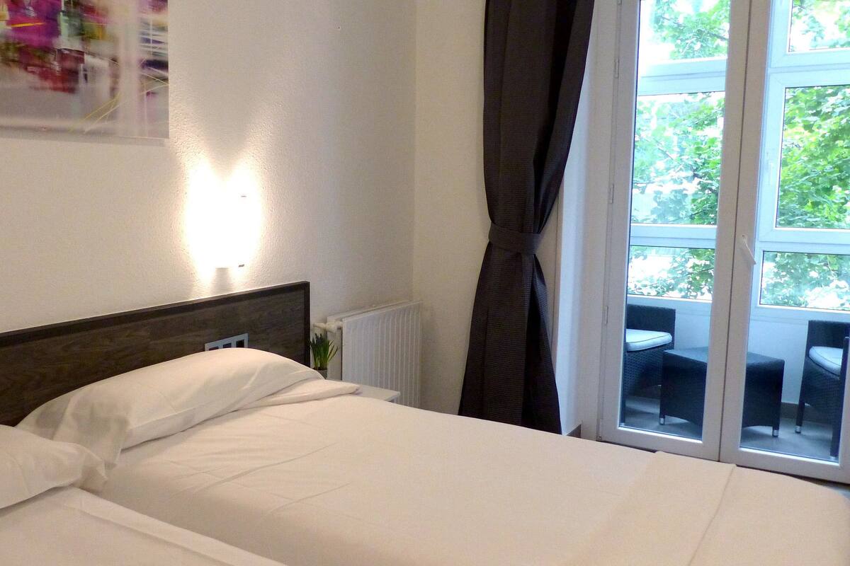 A hotel room with a bed, a radiator, a window with curtains, and a view of greenery outside.