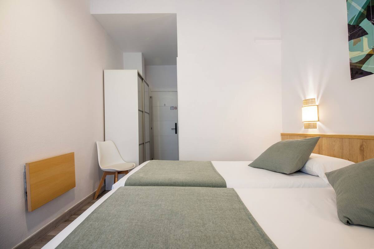 A hotel room with two beds, a chair, a wardrobe, and a wall-mounted light fixture.