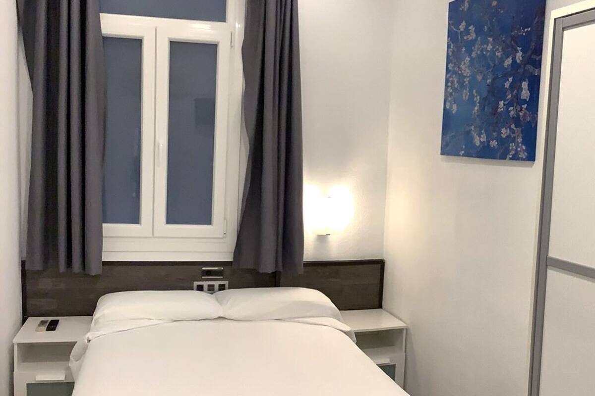 A hotel room with a bed, bedside tables, a nightstand, a window with curtains, and a wall-mounted light.