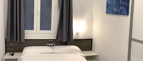Habitación de hotel con cama, mesitas de noche, cortinas en una ventana y una lámpara de pared.
