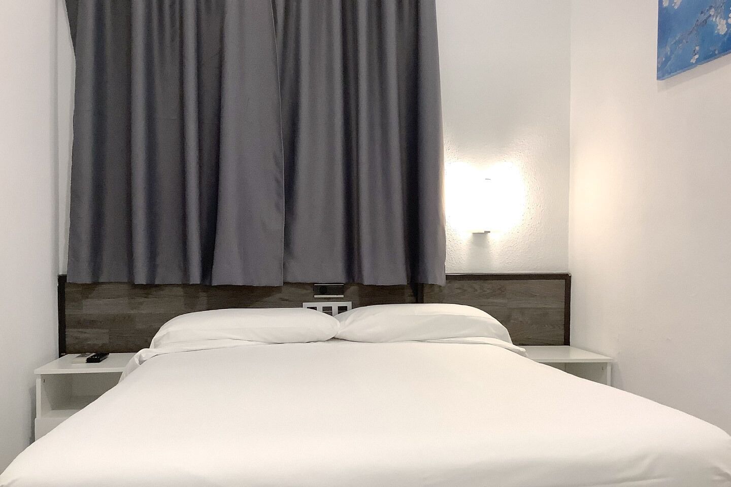 Una habitación de hotel con una cama, ropa de cama blanca, una mesita de noche con un teléfono y una lámpara de pared.