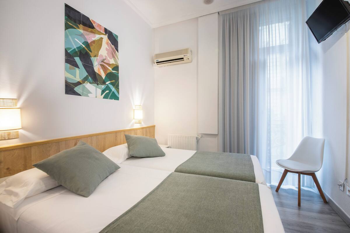 A hotel room with two beds, a large abstract painting, an air conditioning unit, a white chair, and a television mounted on the wall.