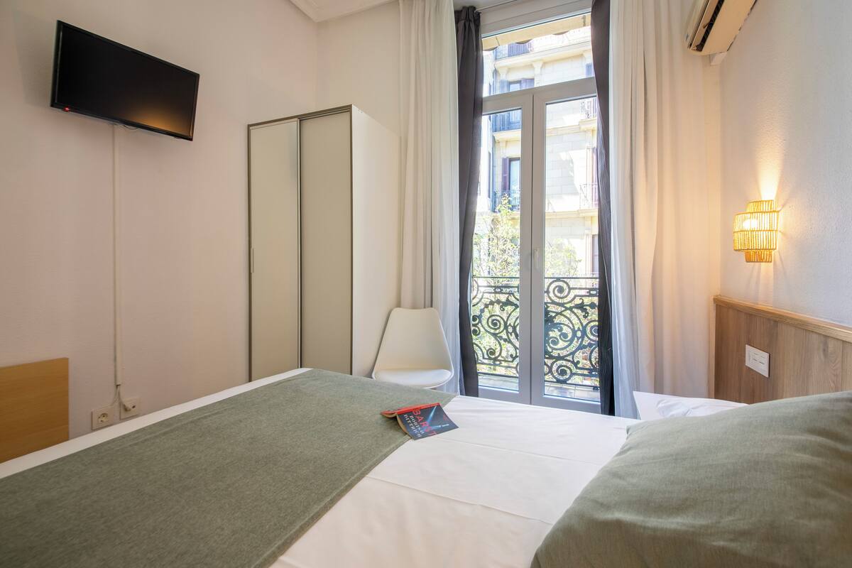 A hotel room with a bed, a TV mounted on the wall, a window with curtains, and a balcony with a railing.