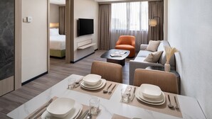 Ein modernes Hotelzimmer mit Essbereich, Wohnbereich mit Sofa und Sesseln, einem Flachbildfernseher und einem großen Fenster mit Vorhängen.