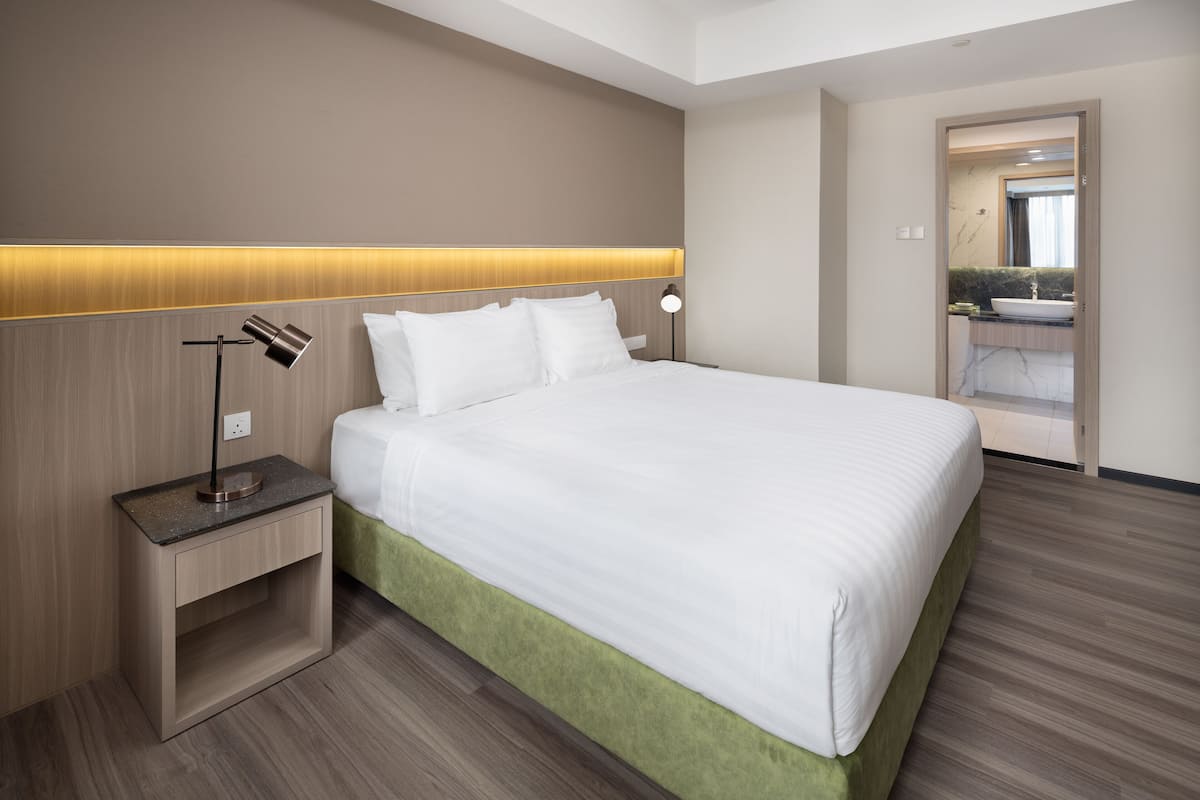 A hotel room with a large bed, a bedside table with a lamp, and a view of the bathroom through an open door.