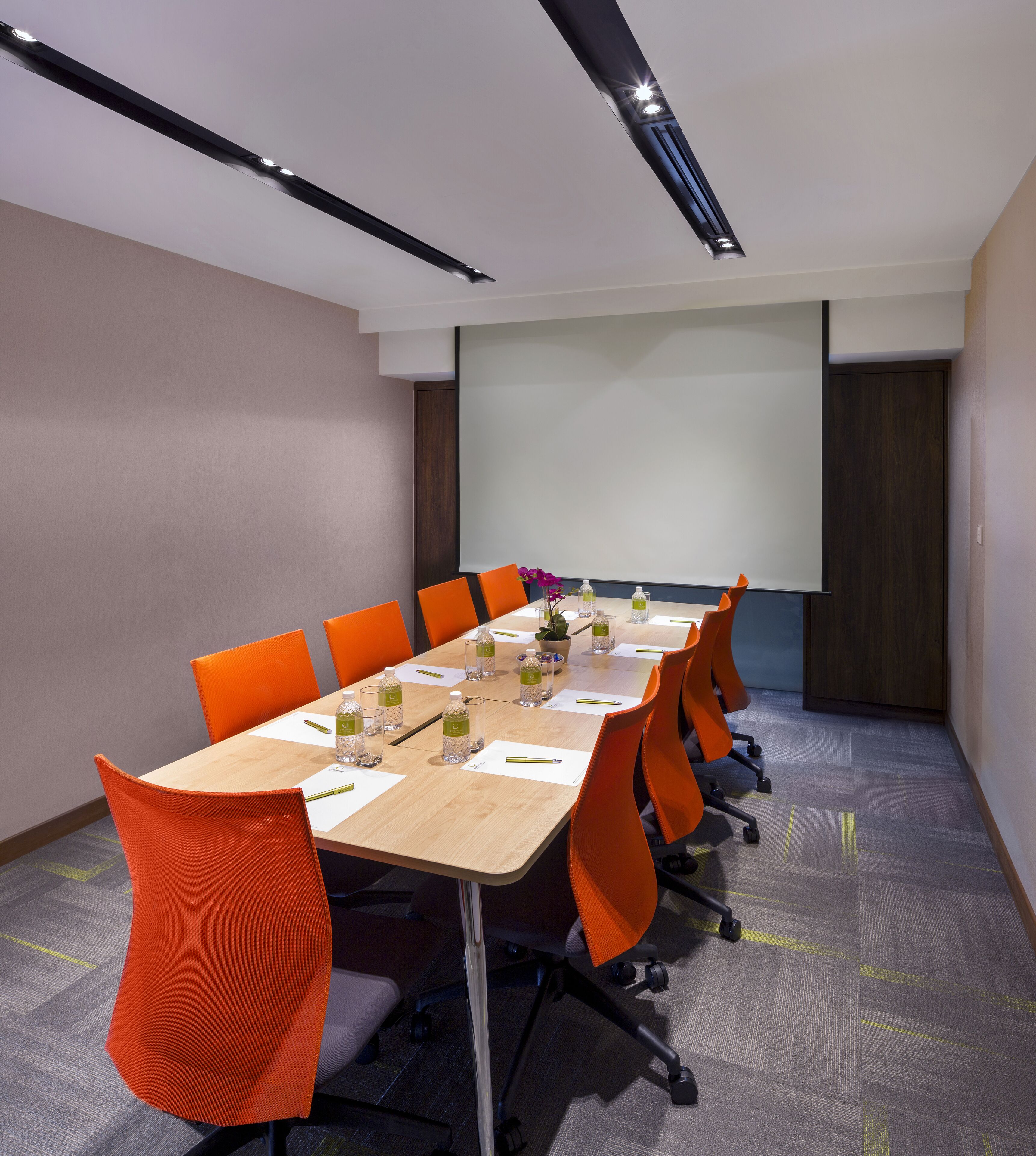 A conference room with a long table, orange chairs, water bottles, and a projector screen.