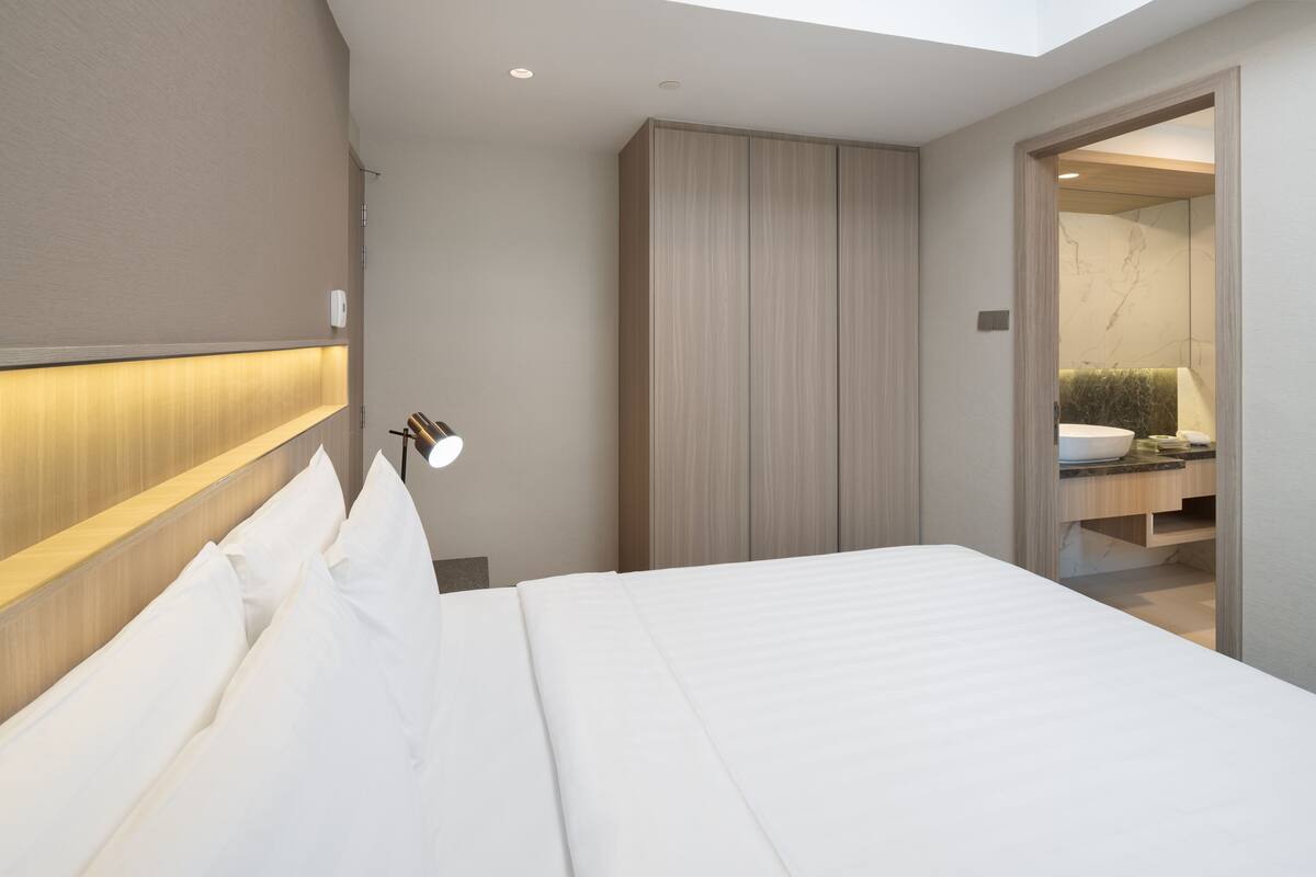 A modern hotel room with a large bed, a bedside lamp, a wardrobe, and a bathroom visible through an open door.