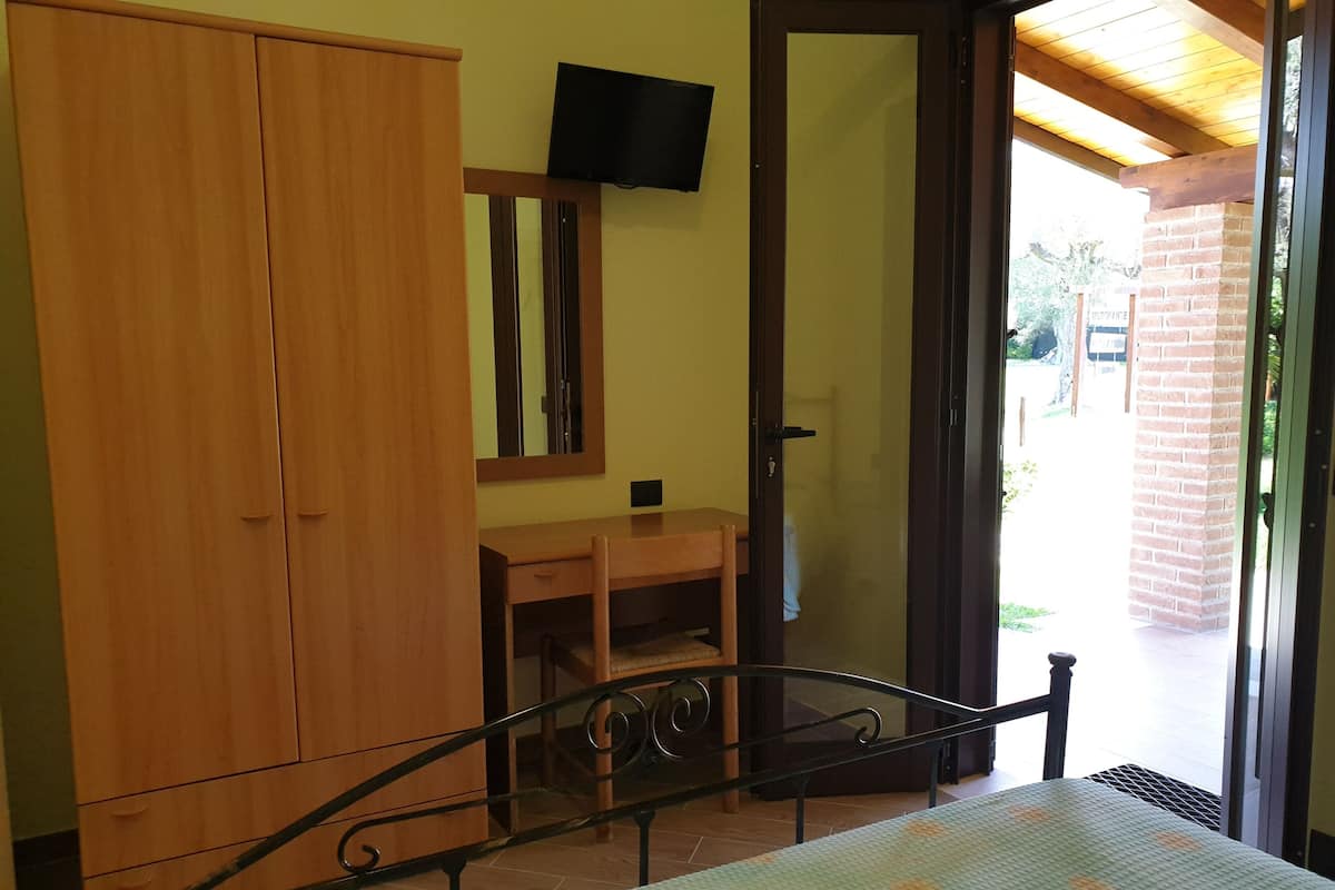 A bedroom with a bed, wooden wardrobe, a small wooden desk, a television mounted on the wall, and a door leading to an outdoor area.