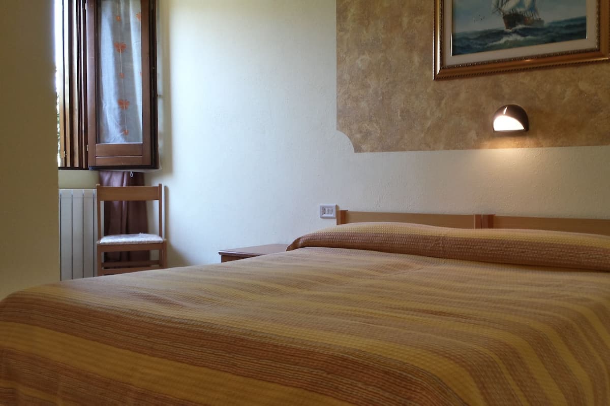 A neatly made bed with a striped bedspread, a nightstand with a lamp, a chair, and a framed picture on the wall.