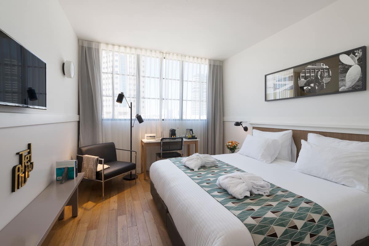 A hotel room with a large bed, a desk with a chair, a flat-screen TV, and a window with curtains.