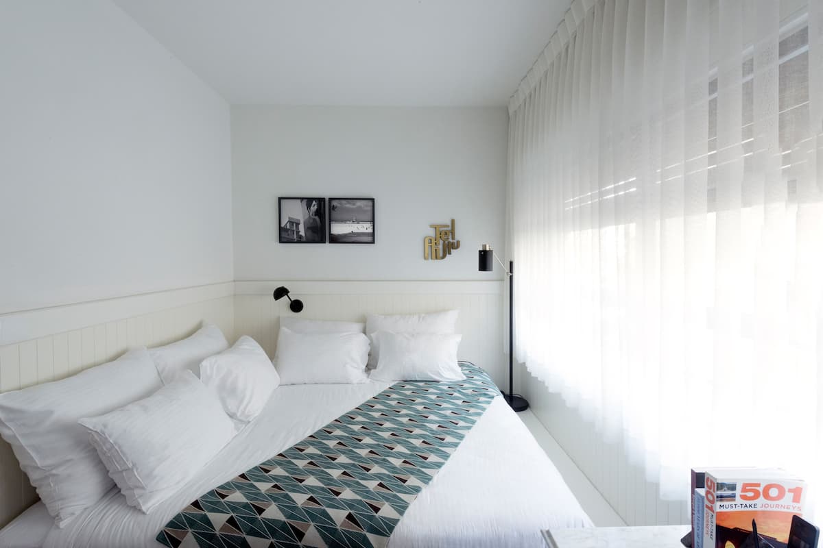 A hotel room with a neatly made bed, a bedside lamp, and a window with sheer curtains.