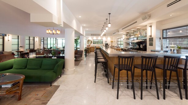 A modern interior with a green sofa, a bar area with stools, and a kitchen in the background. - Lily & Bloom Hotel (Tel Aviv)