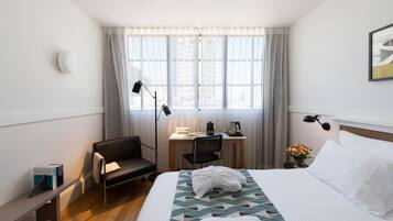 Quarto de hotel com cama, escrivaninha, cadeira, mesa pequena com abajur e janela com cortinas.