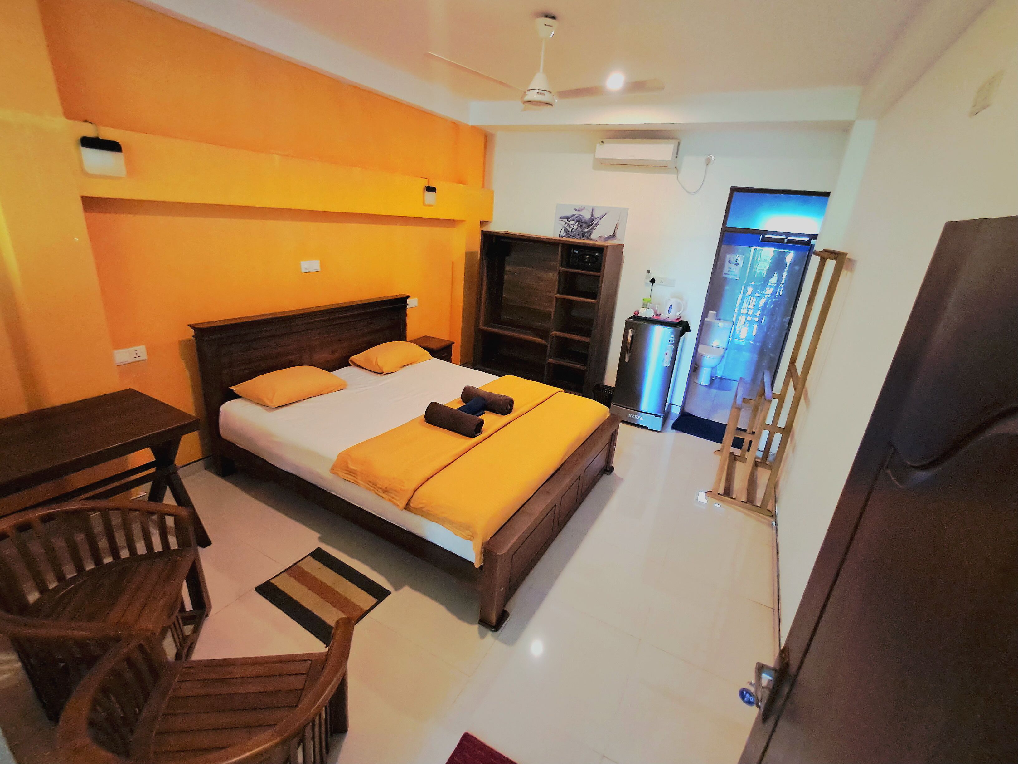 Quarto de hotel com cama, escrivaninha, cadeira, guarda-roupa, pequeno frigorífico e casa de banho visível por uma porta aberta.